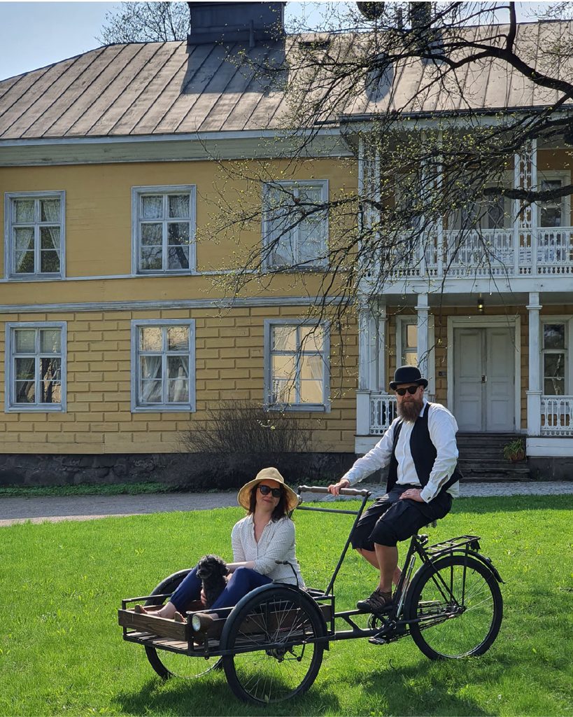 Tavarapolkupyörä aurinkoisena päivänä nurmikolla, jossa mies istuu takana ja nainen edessä ja takana vanha keltainen kartanorakennus.